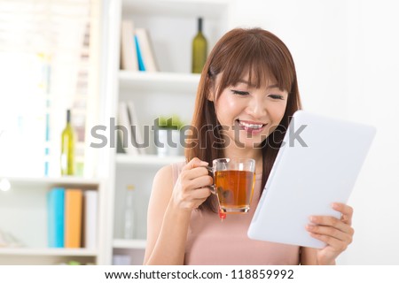 Asian female enjoying cup of tea looking at digital tablet computer inside house