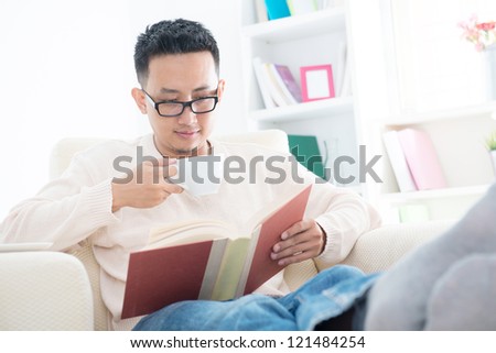 Southeast Asian male reading a book and enjoying a cup of coffee at home, indoor living lifestyle