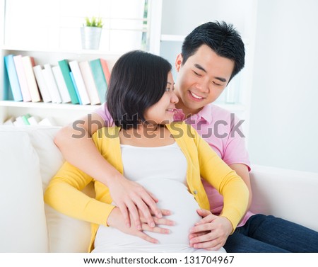 Mom and Dad with hands on the baby, sitting on sofa, Asian pregnant couple living lifestyle.
