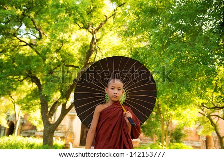 Young Buddhist monk walking outdoors under shade of green tree with umbrella, outside monastery, Myanmar.
