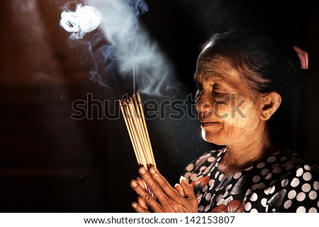 Old wrinkled traditional Asian woman praying with incense sticks inside a temple low light with smoke and beautiful natural sunlight Myanmar Old wrinkled traditional Asian woman praying with incense sticks inside a temple low light with smoke and beautiful natural sunlight Myanmar