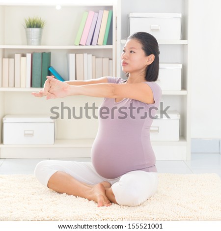 Pregnancy yoga meditation. Full length healthy 8 months pregnant calm Asian woman meditating or doing yoga exercise at home. Relaxation yoga arms stretching pose.