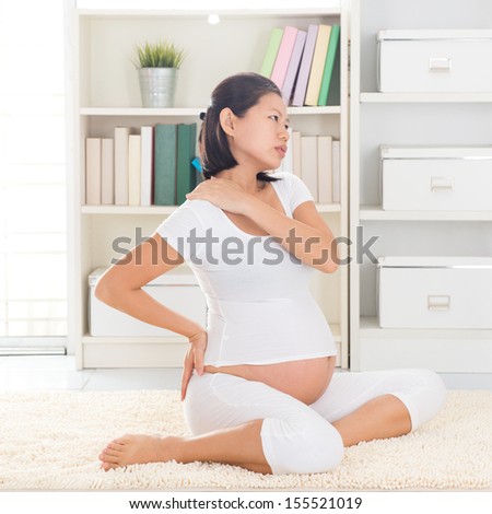Shoulder or neck pain. Eight months pregnant Asian woman holding her back and shoulder while sitting on a floor at home.