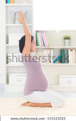 Pregnancy yoga meditation. Full length healthy 8 months pregnant calm Asian woman meditating or doing yoga exercise at home. Relaxation yoga hero pose arms stretching.