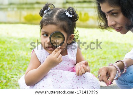 Daughter at outdoor green park with mother. Cute Indian girl peeking through magnifying glass with parent.