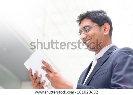 Candid young Asian Indian businessman using digital tablet pc at outdoor building. India male business man, real modern office building as background.