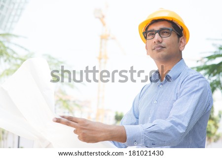 Portrait of a 30s Indian male site contractor engineer with hard hat holding blue print paper at construction site, crane at the background.