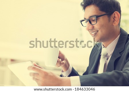 Young Asian Indian businessman using a tablet computer during office break at cafe, relaxing with a cup of coffee. India male business man, in vintage retro style.