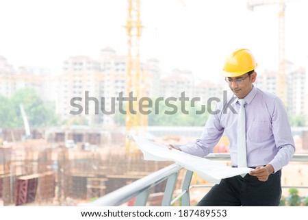 Portrait of a smiling Asian Indian male contractor engineer with hard hat reading blueprint at construction site.
