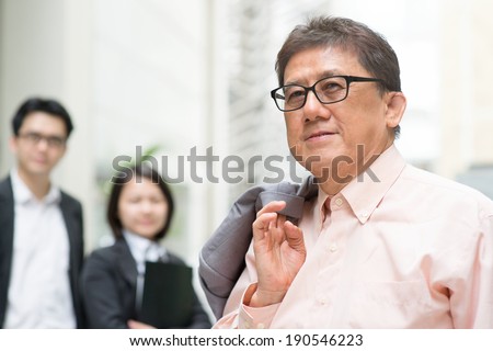 Leader and team member. Portrait of 60s Asian Chinese CEO boss smiling. Senior male businessman and staffs, real modern office building as background.