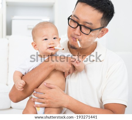 Asian family at home. Bad father smoking with baby together. Cigarette with lit and smoke. Unhealthy lifestyle or stop smoking concept photo.