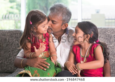Happy Indian family at home. Asian father kissing her daughter, sitting on sofa. Parent and children indoor lifestyle.
