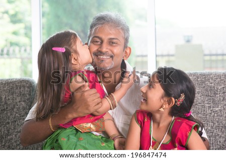 Happy Indian family at home. Asian girl kissing her father, sitting on sofa. Parent and children indoor lifestyle.