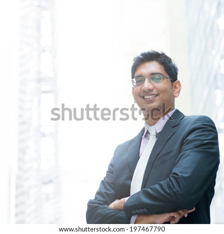 Portrait of a good looking smiling Indian businessman crossed arms standing at modern building, with natural light.