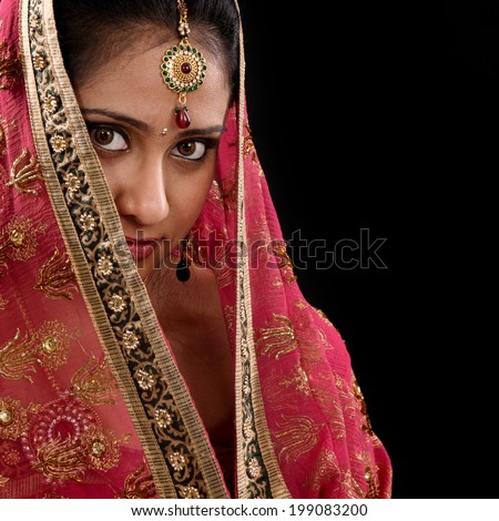 Portrait of beautiful mystery young Indian woman covering her face by headscarf, looking at camera, copy space at side, isolated on black background.