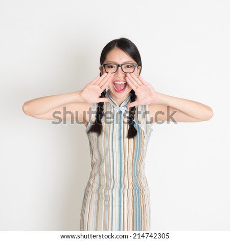 Portrait of Asian Chinese girl  shouting loud, hands next to the mouth, in retro revival style cheongsam standing on plain background.