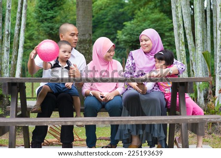 Happy Southeast Asian family sitting at garden bench chatting with each other, outdoor lifestyle at nature green park.