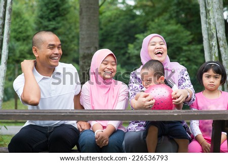 Happy Southeast Asian family sitting at garden bench laughing together, outdoor lifestyle at nature green park.