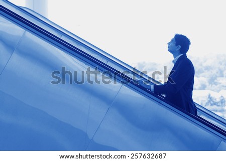 Asian Indian businessman ascending escalator, side view in blue tone.