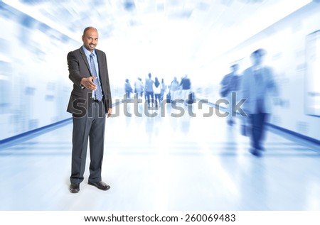 Full body Indian businessman offering hand shake at corridor, inside business building with motion blurred people as background.