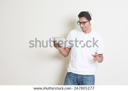 Portrait of handsome Indian guy using tablet pc and enjoying music, standing on plain background with shadow, copy space at side.