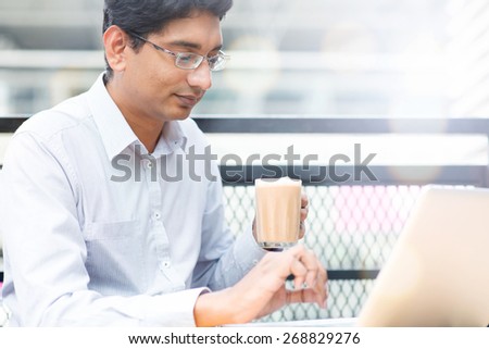 Asian Indian businessman using laptop computer while drinking a cup hot milk tea, outdoor cafe.