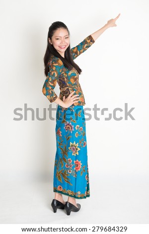 Full body portrait of happy Southeast Asian woman in batik dress finger pointing away, standing on plain background.