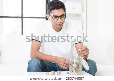 Financial planning concept. Indian guy saving money to glass jar. Asian man sitting on sofa indoor. Handsome male model.