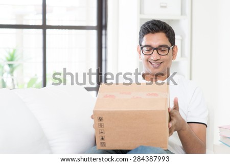 Courier delivery concept. Indian guy received an express parcel and checking the box at home. Asian man sitting on sofa indoor. Handsome male portrait.