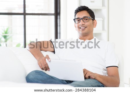 Portrait of Indian guy with digital tablet computer at home. Asian man using internet, relaxed and sitting on sofa indoor. Handsome male model.