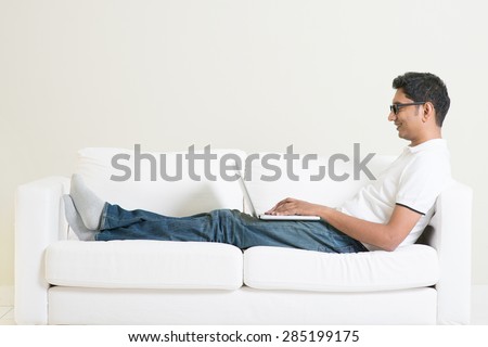 Working from home concept. Indian guy using laptop computer. Asian man relaxed and sitting on sofa indoor. Handsome male model.