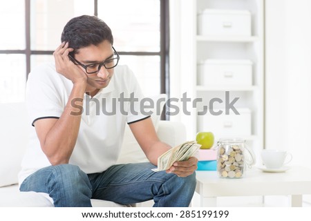 Financial problems concept. Indian guy counting money with sad expression, sitting on sofa at home. Asian male indoor portrait.