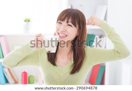 Portrait of happy Asian girl showing strong arm while drinking milk as breakfast. Young woman indoors living lifestyle at home.