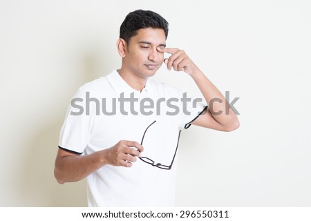 Portrait of Indian guy take off eyeglasses and rubbing his tired eyes. Asian man standing on plain background with shadow and copy space. Handsome male model.