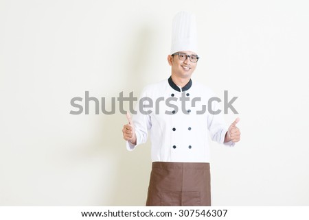 Portrait of handsome Indian male chef in uniform and hat thumbs up and smiling, standing on plain background with shadow, copy space on side.
