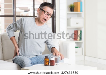 Portrait of casual 50s mature Asian man back pain, pressing on hip with painful expression, sitting on sofa at home, medicines and water on table.