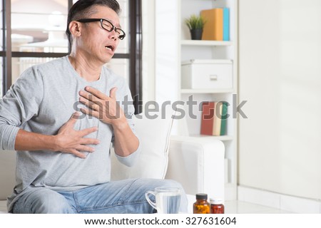 Portrait of casual 50s mature Asian man heartburn, pressing on chest with painful expression, sitting on sofa at home, medicines and water on table.