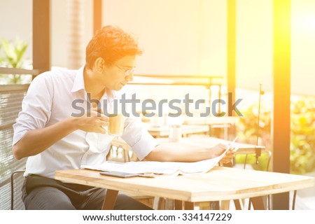 Asian Indian business man reading newspaper while drinking a cup hot milk tea at cafeteria, with beautiful golden sunlight.