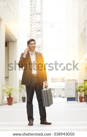 Portrait of fullbody Asian Indian business man using smartphone, standing outside modern office building block, beautiful golden sunlight at background.
