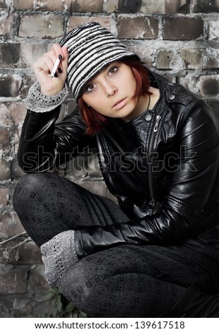 Portrait of young woman smoking cigarette and sitting against a brick old wall