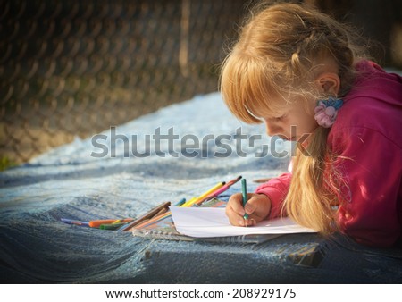 The girl in a red jacket draws a pencil on white paper. She leaned on the boards that are covered with blue tarpaulin.