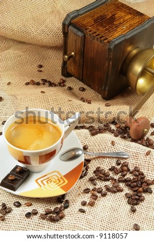 Coffee cup surrounded by coffee grains and coffee-mill in background