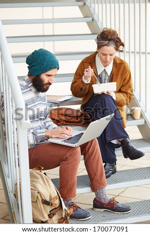 Hipster couple using computer and eating lunch sitting in stairs at university campus