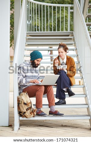 Hipster couple using computer and eating lunch sitting in stairs at university campus