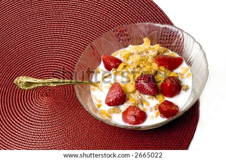 A bowl of cornflakes cereal with whole strawberries,sliced almonds gold fork on a round red straw placemat against a white background. Photographed from above.