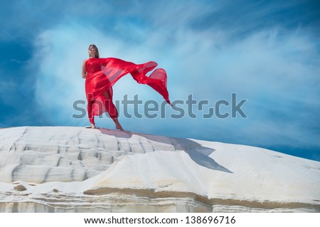Wind blows the red dress on a woman standing against a blue sky.
