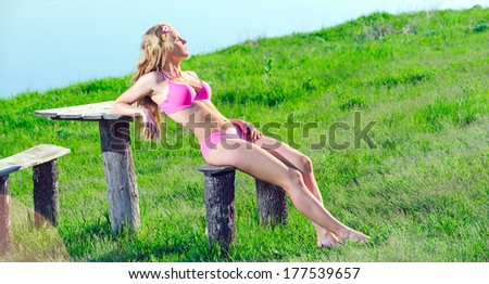 Beautiful positive blond girl clothing in pink swimsuit sit on a wooden bench at the seaside