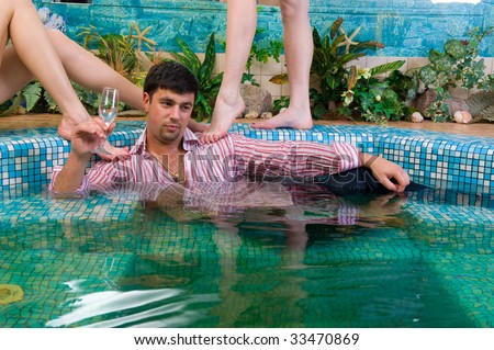 young handsome man with a glass and the clothes in the pool surrounded by women\'s feet