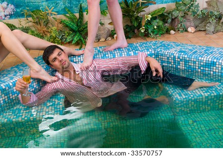young handsome man with a glass and the clothes in the pool surrounded by women\'s feet