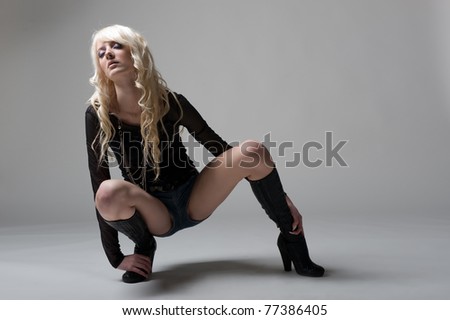 Sexual long-haired blonde girl wearing a jean mini shorts, black shirt and high heels boots posing on the floor in studio on grey background